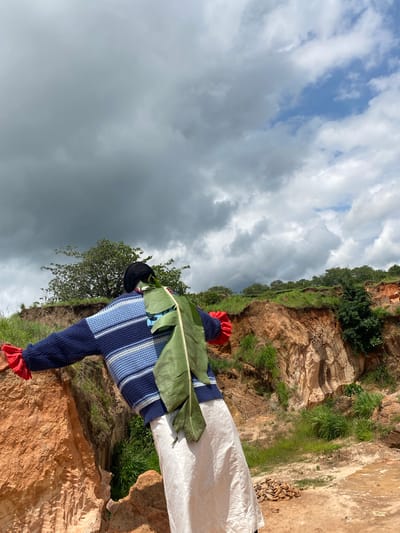 The image shows a person standing in a rural, outdoor environment, likely a quarry or a steep, eroded hillside, in Ufu Esioni, Nigeria. The person is seen from behind, facing away from the camera and towards a dramatic sky filled with both dark, heavy clouds and patches of blue sky. The individual is wearing a blue and white striped sweater, a black head covering, and a long, flowing off-white garment. Red fabric is visible on the cuffs of the sweater, and a large green banana leaf is attached to the back of the sweater. The landscape is characterized by reddish-brown soil, exposed rock faces, and sparse green vegetation. The terrain appears uneven and rugged, suggesting a natural or semi-developed area.