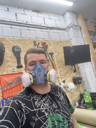 A man wearing a respirator mask and a black t-shirt with white and green graffiti-style writing is taking a selfie in a workshop. The workshop has a white brick wall in the background with exposed pipes and wiring. Various tools are visible on a wooden panel wall, including an angle grinder, a set of wrenches in an orange organizer, a hammer, and a clamp. A small black television is mounted on the wall. A green bottle of lemonade and other items are on a shelf. A digital thermometer on the wall reads 20.8 degrees Celsius.
