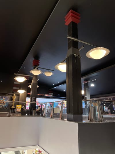 The image shows the interior of a shopping mall in Indooroopilly, Australia, at the coordinates -27.501 latitude and 152.972 longitude. The mall has a black ceiling with several circular light fixtures hanging from it. The light fixtures are attached to rectangular black pillars with red accents. The mall has a white floor, black walls, and a glass railing. Several people are visible in the image. The people are standing and sitting in front of a store. The store has a blue sign and a glass front. There is a gold statue of a person in the background.  There are signs on the wall, one is white with blue letters and the other is a red sign with white letters. The image was likely taken from a higher level in the mall looking down at the floor below.