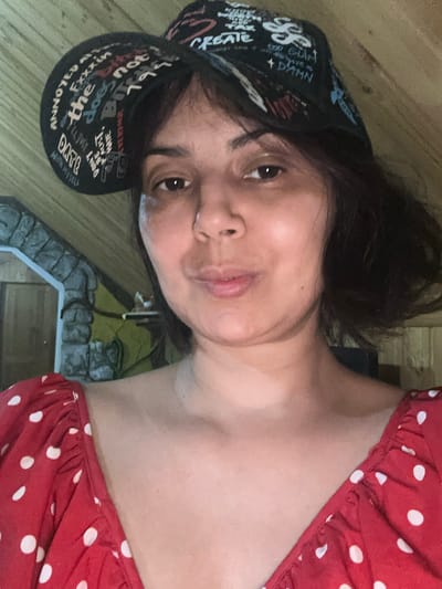 A woman is shown in a close-up, selfie-style shot. She is wearing a black baseball cap with white lettering, and a red dress with white polka dots. The background consists of a wooden ceiling and a stone-like structure. The lighting is warm and appears to be indoors. The specific location in Kalininskaya, Russia is not discernible from the image.