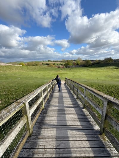 This daytime image captures an expansive outdoor scene in King's Lynn and West Norfolk, United Kingdom. A wooden boardwalk or elevated path with a wooden and wire mesh railing dominates the right side of the frame, extending from the foreground into the mid-ground. A person, dressed in a dark jacket and blue jeans, stands on this path, leaning casually against the railing.
To the left of the path, a vast, verdant green field stretches across the landscape, gently undulating towards the horizon. In the distance, a line of trees and faint structural elements suggest a broader rural setting. The upper left portion of the image showcases a bright blue sky filled with large, fluffy white and grey cumulus clouds, indicating a partially cloudy yet sunny day. The sunlight casts distinct shadows of the railing onto the wooden planks of the path, highlighting the mid-day lighting. The scene evokes a sense of calm and leisure, typical of a natural park or countryside trail. There is no visible text in the image.