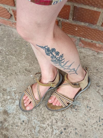 A person's legs, adorned with a tattoo of a sprig of flowers and grass on the left calf, are visible, wearing tan-colored sandals with crisscrossed straps. The individual is standing on a concrete surface, with a brick wall forming the background. The scene is set in Gdov, Russia.