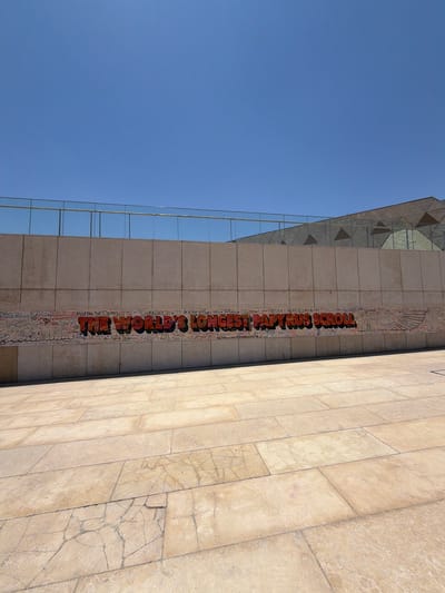The image shows a close-up, worm's-eye view of a building facade with a clear blue sky above. The building features a section of tiled walls, with the tiles appearing to be made of a light-colored stone and showing some cracks. To the left, a modern glass facade reflects the sky.

Dominating the central portion of the tiled wall is a vertical, colorful art installation or mural. Large, three-dimensional orange lettering reads "THE WORLD'S LONGEST PAPYRUS SCROLL." Below this title, the scroll is filled with numerous smaller inscriptions and drawings in various colors, giving the impression of many names, dates, and messages. Some of these smaller inscriptions are legible, including "Katira Cairo," "Ahmet Selim OSMAT HERE 10370204," and "ДАГЕСТАНИС."

The angle suggests the photo was taken looking upwards towards the top of the building, possibly on a sunny day, as indicated by the bright sky and shadows. The setting is likely an urban environment, given the architectural style of the building. The overall impression is one of a public space adorned with a significant artistic display. The presence of so many smaller inscriptions suggests a participatory or commemorative aspect to the "scroll."