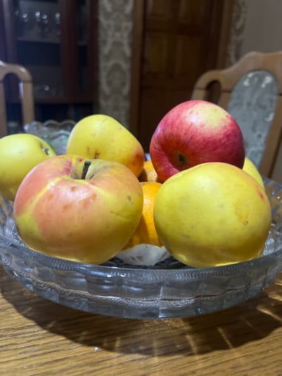 A glass bowl filled with several apples and at least one orange sits on a wooden surface. The apples vary in color, including red and yellow with some showing a mix of both. The bowl is clear and has a textured, possibly decorative, design. The composition is cropped, focusing closely on the fruit and the bowl. The background is out of focus, but suggests an interior space with wooden furniture.