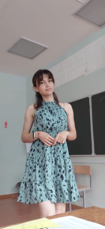 A young woman with brown hair and bangs, wearing a light teal dress with a black abstract pattern, stands in what appears to be a classroom. She has hoop earrings and is wearing rings on her fingers. Her hands are clasped in front of her. A red string bracelet is visible on her left wrist, along with a smartwatch. Behind her, a dark chalkboard is mounted on a light blue wall. Two square ceiling lights are visible. A beige chair with a metal frame is positioned to her right. A small red fish-shaped magnet is on the wall to her left. The environment suggests an indoor setting, likely a school or educational institution, with elements of a classroom visible. The image is taken from a medium-low angle, with the woman as the primary subject.