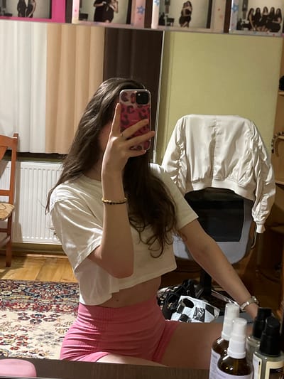 A female individual is depicted taking a mirror selfie within an indoor setting in Popintsi, Bulgaria. Her face is mostly obscured by a smartphone, which has a pink leopard print case and is held in her right hand. She possesses long, dark, wavy hair. She is attired in a white cropped t-shirt and pink form-fitting shorts. A beaded bracelet with dark and amber-colored beads is visible on her left wrist, and a silver ring is present on her right ring finger. A silver-toned watch is worn on her right wrist.
The reflection reveals a room with a light green wall to the right and white and beige vertically striped curtains to the left. A white radiator is positioned below the curtains. The flooring consists of wooden planks, partially covered by a patterned oriental-style rug. A light-colored bomber jacket is draped over the back of a grey office chair to the right. A wooden chair is partially visible on the far left. On the surface holding the mirror, several small bottles, some with spray tops, are arranged in the foreground. A black bag and a black-and-white patterned bag are visible on the floor behind these bottles. Above the main mirror reflection, multiple framed images or photos are visible, depicting individuals and groups of people. Some frames appear pink or have pink decorative elements, including small stars.