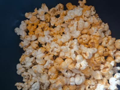 The image shows a close-up, overhead view of a pile of popcorn. The popcorn is a mix of white and golden-yellow kernels, with some kernels appearing more toasted than others. The popcorn is nestled in a dark, possibly black, container. The lighting suggests an indoor setting, and there are no discernible external location cues. The scene is static, with no activity or event taking place beyond the presentation of the popcorn itself. The image is likely intended to be a food photograph, possibly for a menu or advertisement. There is no visible text in the image. The location context of Mineralnye Vody, Russia, does not provide any visual information relevant to the image content.