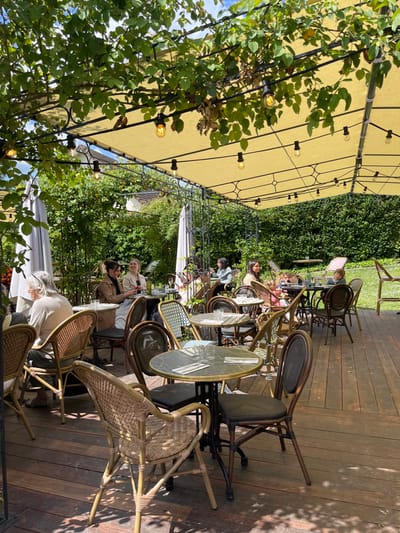 The image shows a patio dining area in the town of Giverny, France. The location is identified by the coordinates 49.075 latitude and 1.536 longitude, indicating the area around the Musée des Impressionnismes Giverny. The patio is covered by a white canvas awning with a black metal frame, supported by wooden beams.  String lights are strung along the frame, adding a festive touch. The patio is surrounded by lush green foliage. There are several groups of people seated at tables and chairs, enjoying their meal. The tables are covered with white tablecloths, and each table has a vase with flowers. The chairs are made of wicker and have a brown finish. The patio floor is made of dark wood, with a decorative pattern.  A wooden railing separates the patio from the lawn beyond. The overall scene is one of relaxed and enjoyable dining outdoors.