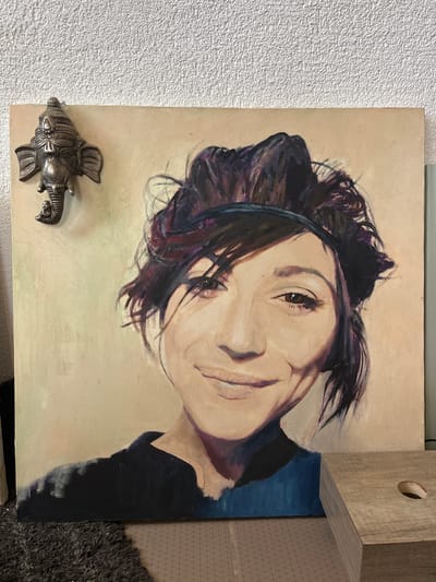 A painting of a woman's face is displayed on an easel against a textured off-white wall. The painting is a close-up portrait, with the woman looking directly at the viewer and smiling. She has dark, messy hair pulled back with a band. Her eyes are dark and expressive, and her lips are slightly parted in a smile. She is wearing a dark top with a blue collar. To the left of the painting, mounted on the wall, is a decorative metal sculpture of an elephant's head. In the foreground, part of a dark, shaggy rug is visible, and to the right, a light-colored wooden object with a circular cutout is placed. The overall lighting suggests an indoor setting. The image does not contain enough information to determine the specific location within Balaklavsky District, Ukraine.