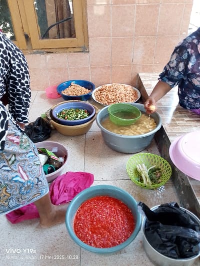 The scene is an outdoor patio in Gegesik, Indonesia (latitude: -6.538, longitude: 108.43). Two women, one with dark and one with light skin, are preparing food. One woman is standing, wearing a black and white polka-dot top and a floral patterned skirt, holding a light-colored vegetable, and appears to be washing it. The other woman, wearing a floral patterned top, is out of frame. In front of them are several bowls, including one filled with water, one with chopped green vegetables, one with diced red pepper, and two with small, orange-colored, chopped pieces. There are two plastic baskets, one green and one beige, one containing a large, green vegetable.  A pink towel lies on the floor next to a large pot with a black plastic bag in it. The environment is tile, a brown, wooden door, and a window.