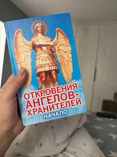 A light-skinned human hand, with a light pink manicure, holds a book vertically in an indoor setting. The book cover is predominantly bright blue, featuring a golden-colored winged angel with a halo, depicted in armor holding an implement. Red Cyrillic text on the cover reads "ОТКРОВЕНИЯ АНГЕЛОВ-ХРАНИТЕЛЕЙ НАЧАЛО," translating to "Revelations of Guardian Angels: Beginning." The hand grasps the left side of the book, with the thumb prominently visible. The subject rests against white bedding adorned with small dark floral or geometric patterns. Behind this, a light blue blanket with white star patterns is visible. A dark, elongated object, possibly a remote control, rests on the star-patterned bedding to the right. The background consists of a plain light grey wall and a blurry brown object, likely furniture. The lighting is soft and even, indicating an interior environment. No discernible visual cues indicate the specific city of Kyzyl-Ozek, Russia.
