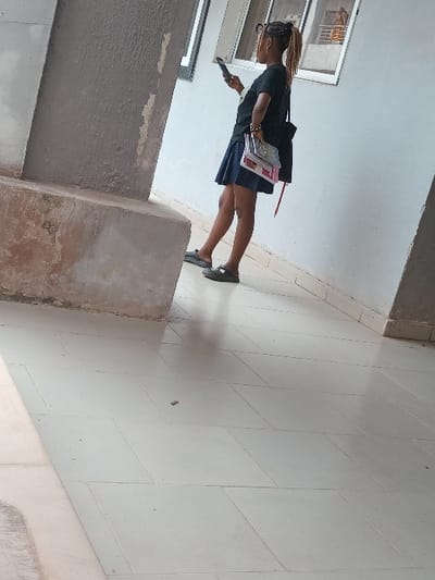 The image captures a female individual standing in a covered walkway of a building in OGBOKO, Nigeria. The subject, observed in right profile, wears a black short-sleeved t-shirt, a dark blue skirt, and dark flat shoes. She has braided hair pulled back and wears eyeglasses. In her left hand, she holds and views a black smartphone. Her right hand carries a stack of approximately four books/notebooks and a dark shoulder bag with a visible red strap. She stands with her weight primarily on her right foot, her left foot slightly forward with the heel raised. The environment consists of a light-colored tiled floor arranged in a diagonal pattern. A light-colored wall with two rectangular, dark-framed windows is visible on the right; one window reflects a metal railing. A thick, textured concrete pillar, light brown-grey in color, occupies the left foreground, showing some minor surface discoloration at its base. The scene is brightly illuminated, suggesting natural light.