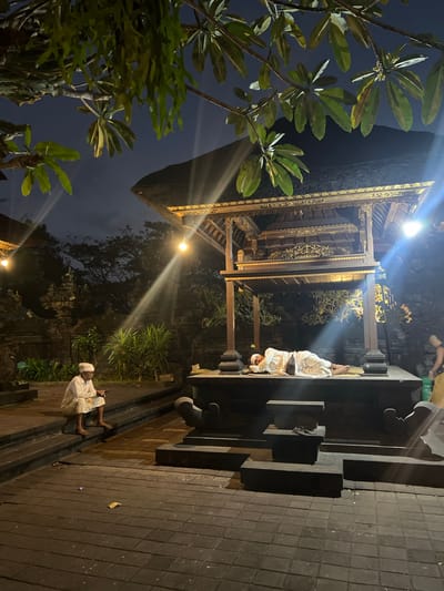 The image depicts an outdoor scene at night, likely in the city of Dawan, Indonesia, possibly at a temple or cultural site given the architecture. In the foreground, a man dressed in white traditional clothing, including a head covering, is seated on stone steps, looking down at a smartphone he holds in his hands. He appears to be smoking a cigarette. To the right, a similar man, also in white traditional attire, is lying down on a raised platform, seemingly asleep. He is covered by white fabric and is positioned with his head resting on a dark pillow. The background shows intricately carved wooden structures with a thatched roof, illuminated by warm lighting. Lush green plants and trees are visible, some of which overhang the scene from above. The ground is paved with dark square tiles. The sky is dark blue, indicating nighttime. There is a hint of another person in the far right background, partially obscured.