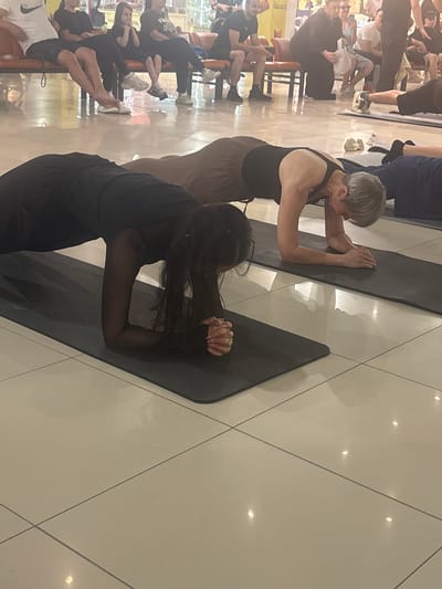 In Tri Protoka, Russia, two women are performing planks on yoga mats on a tiled floor in what appears to be an indoor public space, possibly a community center or a mall. The woman in the foreground on the left is wearing a black outfit and has long, dark hair. The woman on the right is wearing a dark tank top and brown pants and has short, gray hair. In the background, several other people are seated on a bench. One person is wearing a white t-shirt and dark shorts, another is wearing a black t-shirt and dark pants, and a third is wearing a black dress. The environment is well-lit, with some reflections on the tiled floor suggesting overhead lighting.
