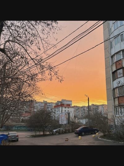 The image depicts an urban landscape with a dramatic orange sky. In the foreground, a bare-branched tree is on the left, and a multi-story building with balconies is on the right. Power lines crisscross the sky. Several apartment buildings are visible in the middle ground, with some trees interspersed. Cars are parked on the asphalt pavement in front of the buildings. There are no identifiable people in the image. The scene appears to be an exterior view, possibly a residential area, with the setting sun casting a warm glow. The location is not Saint-Août, France.