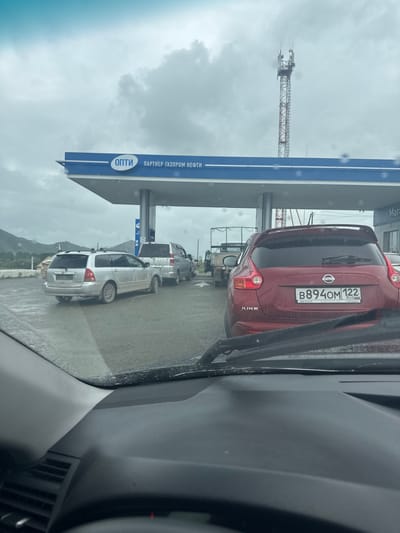 The image shows a line of cars at a gas station. The gas station canopy has blue and white signage, with the words "ОПТИ" and "ПАРТНЕР ГАЗПРОМ НЕФТИ" visible. In the foreground, the interior of a car is visible, including the dashboard and windshield with rain drops. A red Nissan Juke is the first car in line. Behind it, a silver station wagon and a silver minivan are visible. A cell tower is visible in the background against a cloudy sky. The scene appears to be a typical gas station in Ust-Kan, Russia, given the Cyrillic signage.