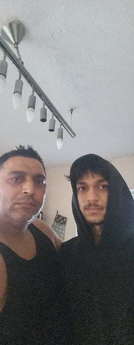 The image displays two males inside an indoor environment. The specific location in Gloucester, United Kingdom, cannot be determined from the visual information provided. The male on the left is of apparent middle age, with dark, spiked hair and facial stubble, wearing a black tank top. The male on the right is younger, with dark hair, a light mustache and goatee, wearing a black hooded sweatshirt with the hood raised. Both individuals are positioned facing forward, looking directly towards the viewer. Above them, a silver-toned, linear track light fixture with multiple bulb holders, some containing white-tipped bulbs, is affixed to a white, textured ceiling. A partially obscured, patterned object is visible in the background behind the younger male's right shoulder. The room features light-colored walls and appears to be illuminated by artificial overhead light.
