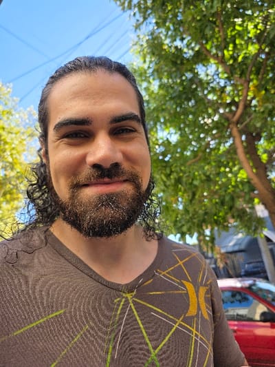 The image displays an outdoor, brightly lit, close-up shot of a single male subject in Villa San Andrés, Argentina. The subject, a man with dark curly hair pulled back, a full dark beard, and a mustache, is centered and smiling directly at the camera. He is wearing a short-sleeved, brown V-neck t-shirt patterned with intersecting thin lines in yellow, white, and light green, and a faded yellow-orange Hurley logo on the right chest area. The background is out of focus. The upper left portion shows a clear blue sky with two thin, dark power lines crossing diagonally. Green foliage from trees is visible to the subject's left and prominently on the right side of the background. A red vehicle is partially discernible in the blurred far background on the right, positioned near a dark, indistinct building or structure. The scene is illuminated by natural daylight.