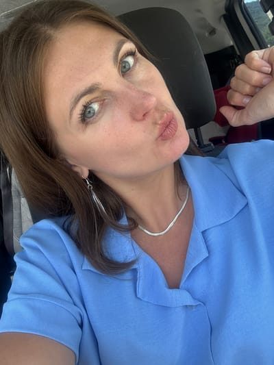 A close-up, upside-down selfie captures a woman's face with her lips pursed in a kiss. She is wearing a light blue collared shirt and a silver necklace. A long, silver, feather-like earring dangles from her left ear. Her blue eyes are wide and looking directly at the camera. Her skin has a light dusting of freckles across her nose and cheeks. Her brown hair is styled down and frames her face. In the background, part of a black car seat is visible. To the left of the woman's head, a hand is holding a piece of dark red fabric. The lighting suggests it is daytime, likely outdoors, given the hints of greenery in the upper left corner of the original image. The overall tone is playful and self-aware.