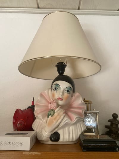 The image shows a close-up of a shelf displaying several decorative items. The central object is a ceramic lamp with a lampshade, featuring a stylized clown's face and torso. The clown has theatrical makeup, with blue eyeshadow, rouged cheeks, and a pink rose held in its hands. To the left of the lamp is a red ceramic pig figurine with a smiling face. In front of the pig is a white box with various icons printed on it, possibly representing product features. To the right of the lamp, there is a small glass-cased clock with visible gears, and behind it, a tiered decorative item, possibly made of wood or metal. The shelf itself appears to be made of wood, with a textured white wall in the background. The lighting suggests an indoor setting. There are no people or discernible actions captured in this static scene. There is no information in the image to suggest a location in the city of Kassel, Germany.