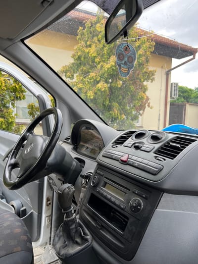 The image shows the interior of a Mercedes-Benz Vito van, parked outdoors in Žatčany, Czechia. The vehicle's interior is visible from the driver's perspective, looking towards the dashboard, steering wheel, and windshield. A decorative sugar skull air freshener hangs from the rearview mirror. Outside the vehicle, a house with a red-tiled roof and green trees is visible through the windshield. An air conditioning unit is mounted on the exterior wall of the house. The scene appears to be a residential area.