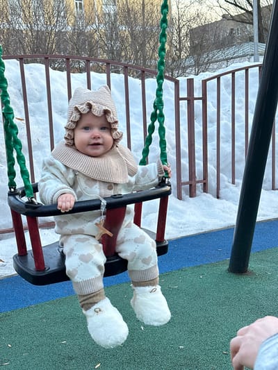 An infant is seated in a playground swing in Sadovaya, Russia. The infant wears a light beige knitted bonnet with ruffled edges, a matching knitted bib, a cream-colored cardigan with light brown buttons, and cream-colored pants adorned with light brown heart patterns. White, fluffy booties with small gold-colored ring embellishments are on the infant's feet. A small wooden cross pendant hangs from a clear cord attached to the front restraint bar of the black and red plastic swing. The infant looks forward with a smile. The swing's ropes are green and twisted. The ground beneath the swing is a blue and green speckled rubber safety surface. In the background, a brown metal fence separates the playground from deep snowdrifts. Bare trees and residential buildings are visible beyond the snow. A partial human hand is visible in the lower right foreground.