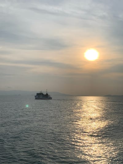The photograph depicts a seascape with a hazy sky dominated by a bright sun. Light reflects off the water's surface, creating a shimmering effect. A ferry boat is visible in the middle ground, traversing the water. Hazy land is on the left and far right. There is no definitive evidence to confirm it is Thong Nian, Thailand. No people are visible.