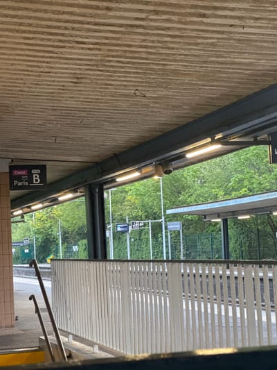 This is an eye-level photograph taken at the Champigny-sur-Marne train station in France. The photo appears to have been taken at the station, with a view of the platform and greenery visible through the station's structures. A sign indicates the direction "Ouest" (West) and "Paris" for line B. A gray metal structure with linear lights runs along the top left of the frame, adjacent to a textured wall. The right side of the photo shows part of the platform with a covered walkway. Beyond the platform, trees and shrubbery are visible, along with signs with numbers on them.