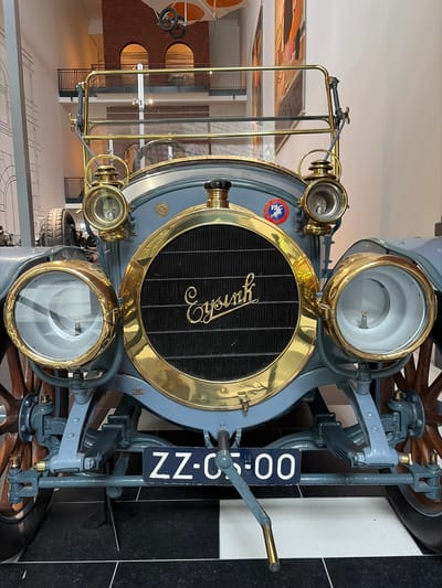 An antique automobile, oriented vertically, is prominently displayed indoors, likely in a museum setting in Wassenaar, Netherlands. The front of the vintage car, painted in a muted grey-blue, is the primary subject. Its most striking features are the polished brass components: a large, circular radiator surround, two prominent main headlights, and two smaller auxiliary lights positioned below. The central radiator grille, a black mesh, bears the car's name "Spyker" in elegant gold script.
A round badge affixed to the left side of the radiator displays the text "PAC AUTOMOBILES" surrounding a red and blue emblem. The car's front suspension system, also in grey-blue, is visible, along with parts of a wooden spoked wheel on the right. A dark blue license plate with white characters, reading "ZZ-C5-00", is mounted vertically on the right side of the front assembly. A brass-tipped hand crank extends from the lower right.
The background reveals a spacious, well-lit interior with a blend of modern and industrial architectural elements. Reflections suggest glass barriers or polished surfaces. Further back, textured walls, possibly exposed brick, feature large circular openings, while a patterned floor (black and white squares near the car) transitions to a lighter color. The car appears stationary and is the sole focus of the scene, with no people visible. The setting suggests an exhibition or permanent display of historical vehicles.