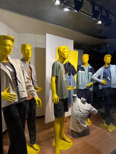 A store window display in Villafranca di Verona, Italy, features five horizontally arranged mannequins. All are painted a bright yellow color, including hair and feet, and are positioned on shelves against a wall. The mannequins are dressed in casual clothing with visible price tags, suggesting a fashion retail setting. The background reveals a reflection of outdoor features like lights, adding depth and indication that the scene is being viewed from a store exterior. The general atmosphere is modern.