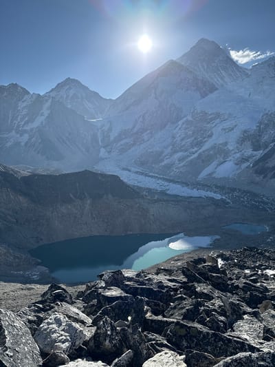 This media file captures a majestic, high-altitude mountain landscape, looking down from a steep, rocky vantage point. The scene is dominated by massive, snow-capped peaks that rise dramatically on the left and center, their rugged faces showing both white snowfields and exposed dark rock. These mountains recede in layers, conveying immense scale and depth.
To the right and below the towering peaks, several serene glacial lakes or tarns are visible. Their waters exhibit a striking turquoise color, reflecting the clear blue sky. One of the lakes appears partially covered by ice or snow along its edge.
The immediate foreground on the right is composed of a dark, sharply textured, and steeply angled rocky slope, suggesting the photographer is positioned on a high, craggy ridge or cliff edge overlooking the valley and lakes.
The sky, visible in the upper left, is a vibrant clear blue. A bright, dazzling sun is prominently featured in the upper-left quadrant, casting a strong glare and producing a distinct lens flare, indicating intense daylight, likely mid-morning or early afternoon. A few wispy white clouds are scattered near the mountain summits.
The overall impression is one of immense natural grandeur, remoteness, and pristine beauty, characteristic of an alpine environment like the Himalayas, as suggested by the Khumjung, Nepal context. There is no visible human activity or text in the image.