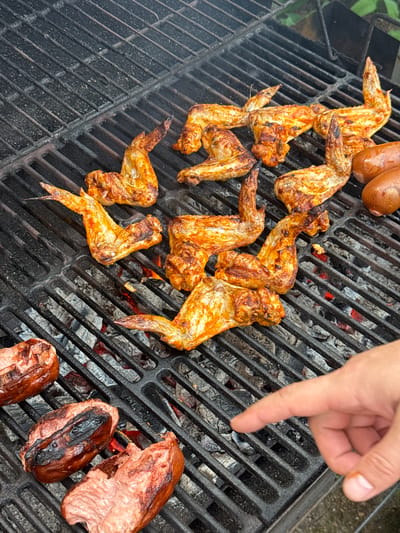 In Усть-Мана, Russia, a close-up, high-angle shot captures chicken wings and sausages grilling on a charcoal grill. The grill grates are dark and visibly used, with visible charcoal embers glowing red beneath. Several chicken wings, some appearing seasoned with a reddish spice, are arranged on the upper left and center of the grill. To the right of the wings, two browned sausages are positioned. On the lower left, three more sausages are visible, one split open revealing its raw interior. A human hand with a visible thumb and index finger extends from the bottom right corner of the frame, with the index finger pointing towards the grill. The background is out of focus, suggesting an outdoor setting.