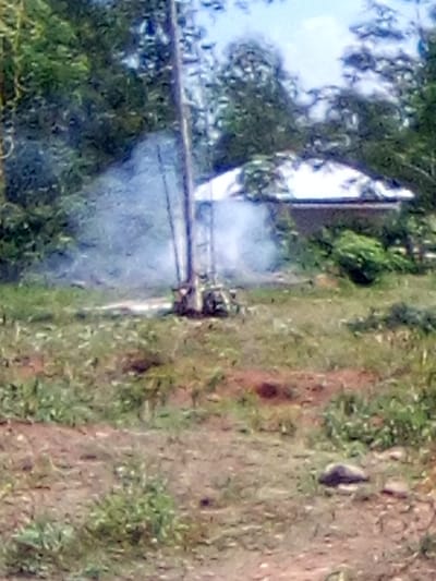 The image depicts an outdoor scene in West Sakwa ward, Kenya. In the midground, a vertical dark-colored pole or structure stands, possibly associated with a piece of machinery or equipment located at its base. This equipment is actively emitting a significant plume of white smoke or steam that disperses upwards and to the left. The ground directly surrounding the equipment appears disturbed and reddish-brown, contrasting with the surrounding sparse green grass. In the background, partially obscured by green foliage, a building with a light-colored, rounded or dome-shaped roof is visible. Dense green trees and bushes frame the upper portions of the image, with a light blue sky visible above them on the right. The foreground consists of dry, sparsely vegetated ground with some small rocks. No individuals are visible in the scene.