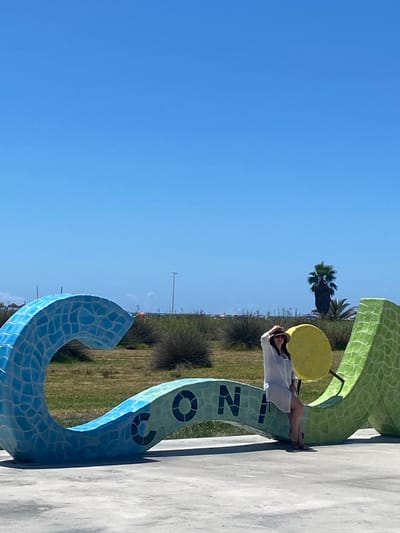 A woman wearing a white cover-up and a straw hat poses for a photo in front of a large, colorful, mosaic sculpture that spells out "CONIL". The sculpture is a vibrant blue on the left, transitioning to light blue and green, with the letters "CONIL" visible in dark blue. To the right of the letters, there's a yellow circular element and a green serpentine shape. The woman is leaning against the sculpture, her hand on her hat, looking towards the camera. The background consists of dry vegetation, a palm tree, and a clear blue sky. In the distance, there are hints of buildings and infrastructure, possibly a road or coastline, suggesting an outdoor, possibly coastal, setting. The ground in the foreground is a paved surface. The overall scene conveys a bright, sunny day in a public outdoor space, likely a promenade or park in Conil De La Frontera, Spain, given the sculpture's lettering.