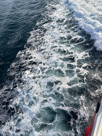 The scene depicts a detailed view of turbulent seawater, characteristic of a vessel's wake, located in the waters off Kamariotissa, Greece. The upper left portion of the image displays relatively calm, dark blue-green sea. A dynamic transition occurs towards the center and right, where the water is heavily aerated, forming an expansive pattern of bright white foam and churning, lighter blue-green water. This foam consists of numerous interconnected bubbles and swirling patterns. In the lower right corner, a segment of a white ship's hull with a distinct red stripe is visible, indicating the source of the generated wake. No individuals, landmasses, or other specific objects beyond the water and ship hull are present in the frame.