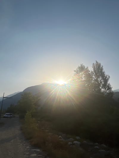 The image captures a sunburst effect behind silhouetted trees and mountains, possibly in the vicinity of Benasque, Spain. A gravel path curves into the foreground on the left, where a white van is parked. The sky is a pale blue, fading to white near the horizon, with the sun's rays creating a rainbow effect as it peeks over a mountain range. The surrounding landscape is dominated by dense, dark foliage and rocky terrain.