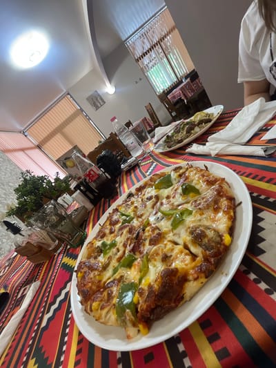 The image shows a dining table with a tablecloth featuring a geometric pattern with red, yellow, and dark blue stripes. A large oval white plate contains a pizza with visible toppings including cheese, green bell peppers, and corn. A second, smaller oval plate holds another dish. Silverware and white napkins are present. Behind the food items are bottles and other objects. The room includes a window with blinds, and a glimpse of the interior suggests a residential setting. There is no verifiable information that locates this image in Dolna Mitropolia, Bulgaria.