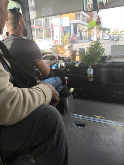 Inside a vehicle, likely a bus, a man in a black t-shirt drives. The dashboard is visible, with cups, a plant, and a water bottle. A passenger, partially shown, wears a light-colored sweater and dark pants. Through the windshield, a street with buildings, cars, and pedestrians can be seen in daylight. The overall setting appears to be an urban environment during the day.