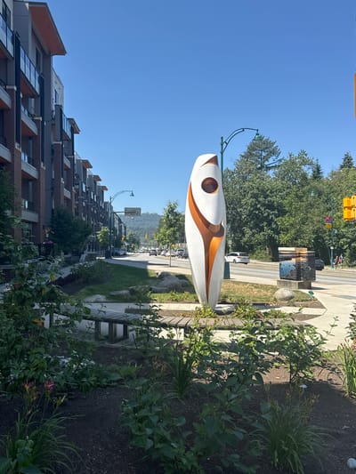 This is a photograph of a street-level view in Port Moody, Canada, likely taken during daytime. On the left side of the image, a modern brick and glass building complex with multiple stories is visible, featuring balconies with glass railings. A paved sidewalk runs in front of the building, bordered by a landscaped area with a wooden bench and various plants, including flowering plants and green bushes.
In the foreground, a large, abstract white and orange sculpture stands prominently. Behind the sculpture, a street with moderate traffic is visible, with cars and streetlights lining the road. A number of trees with lush green foliage are situated along the street and in the background, with a treed hill visible in the distance. To the right of the sculpture, a concrete pedestal supports two rectangular objects, possibly art installations or utility boxes, adorned with images of water and landscapes. Further to the right, a traffic signal with yellow and red lights is present. Two individuals are seen walking on the sidewalk in the distance. The sky is clear and blue.