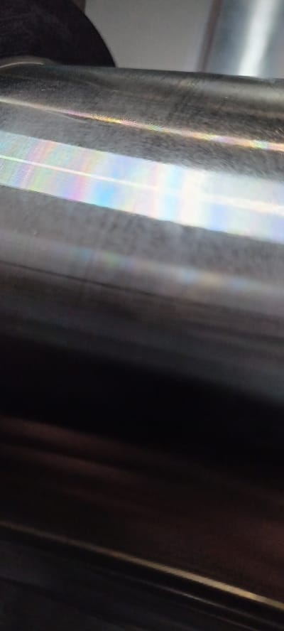 The image is a close-up, abstract shot of a smooth, dark, reflective surface, possibly metal or polished plastic. There are bands of iridescent colors, resembling a rainbow or oil slick effect, streaking across the upper portion of the surface. These colors are likely caused by light refraction or interference. The lower portion of the image is almost entirely black, with subtle variations in tone suggesting a curved or glossy texture. No specific location in Kiselyovsk, Russia, can be identified from this abstract imagery.