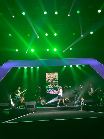 The image shows a stage setup with a performer lying on the stage floor, possibly singing or performing. There are band members on either side of the performer, playing instruments. A screen with a floral design is visible behind the performer. The stage is brightly lit with green stage lights, and there's a geometric frame around the central stage area, illuminated with blue and purple lights. Equipment like drums and speakers is also visible. The image was taken at Salembaran, Indonesia.