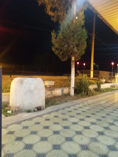 The image depicts a nighttime scene in an outdoor urban setting. In the foreground, a patterned paved walkway, composed of light-colored hexagonal tiles interspersed with darker square tiles, leads into the composition. To the left of the walkway, a large, roughly cylindrical white object with a rounded top is partially visible. Beyond this object, grass and some foliage are present.
A slender tree with a white trunk stands to the left of center, its branches extending upwards. Behind the tree, a yellow pole supports a light fixture, and a low railing with vertical supports is visible. Several small, red-glowing lights are mounted on poles in the middle ground, spaced out along the railing. A structure with a yellow roof is visible in the upper right corner of the image, casting a warm light. The background is dark, indicating nighttime, with silhouetted trees and possibly distant structures. The overall impression is of a public area, possibly a park or a square, illuminated by artificial lights. The location is identified as Kyzyl-Kyya, Kyrgyzstan.