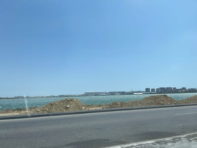 The media file depicts a wide, elevated view of a body of water with buildings in the distance. A paved road occupies the foreground, with a curb and a strip of dry earth and vegetation separating it from the water. Two large mounds of soil are present in the mid-ground, positioned along the edge of the water. The water itself appears somewhat choppy, suggesting wind.

Across the water, a cityscape is visible, characterized by numerous buildings of varying heights, including what appears to be a stadium or large arena with a white, curved roof, and a suspension bridge. The sky is clear and a bright blue, indicating fair weather and likely daytime, possibly late morning or afternoon given the lighting. The overall setting suggests an urban or semi-urban environment adjacent to a large body of water, possibly a bay or lake, in Sabunchu Raion, Azerbaijan. No people or specific activities are discernible. The scene conveys a sense of openness and a coastal or lakeside landscape. There is no visible text in the image.