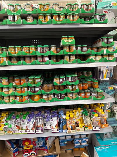 This image captures a well-stocked aisle in a retail store, primarily featuring baby food and related products. The scene is dominated by numerous glass jars of baby food, predominantly the "Beech-Nut" brand, neatly arranged in green cardboard trays on multiple shelves. These shelves appear horizontally due to the image being rotated 90 degrees clockwise.

To the right of the jarred baby food, a section displays various baby food pouches, including brands like "SUPER morning" (from Happy Baby Organics) and "munchkin," which are presented upright in their packaging. Further right, a diverse selection of baby-related items is visible, such as packaged baby feeding spoons (one package shows "OXO" branding), small cups, and what appears to be a "nutribullet BABY" blender set in its box. Several boxes labeled "KID" containing toy vehicles are also present on a lower shelf. The shelves are full, indicating a well-maintained stock.