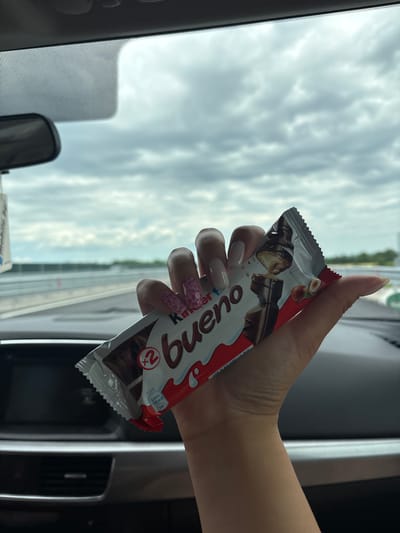 A person is holding a "Kinder Bueno" chocolate bar in their hand inside a car. The background shows a cloudy sky visible through the car window and a road or overpass. It is difficult to ascertain the location within the city of Peshterna, Bulgaria, without additional information.