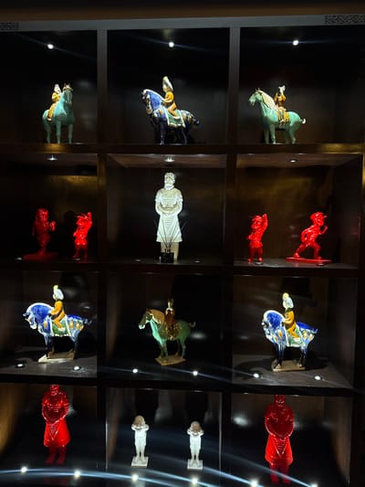 This image displays a collection of ceramic figurines arranged in a multi-shelf display cabinet. The cabinet is divided into nine cubbies, each housing one or more figurines. The figurines depict various subjects, including horses with riders, standing figures, and a single large warrior figure. The overall aesthetic suggests a display of decorative or historical art pieces. The setting appears to be an indoor environment, likely a museum or a private collection, with subtle lighting illuminating the individual pieces. The image does not provide any information that would allow for identification of the city as Barvikha, Russia.