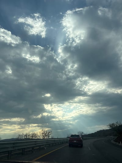 A red car travels along a highway, the passenger's perspective captures the sky, filled with layers of dark and bright clouds. Sunlight streams through the clouds. A metal guardrail runs alongside the road. Trees and hills are visible beyond the guardrail. The location is unknown. The image lacks the necessary information to identify the city or neighborhood. No people or actions are visible.