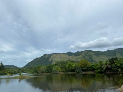 The image depicts a serene landscape featuring a body of water reflecting the surrounding environment. In the foreground, the calm water surface is marked by gentle ripples, suggesting a tranquil atmosphere. Across the water, a lush, green shoreline is densely populated with various trees, creating a verdant barrier. Beyond the trees, a majestic mountain range rises, its slopes covered in a mix of greenery and grassy patches. The sky above is overcast, with scattered clouds contributing to a soft, diffused lighting. The scene appears to be a natural setting, possibly a lake or river, with the mountainous backdrop indicating a location with significant elevation. The presence of palm trees on the left side of the image suggests a tropical or subtropical climate. The overall impression is one of natural beauty and tranquility. The location is identified as Aneuk Paya, Indonesia.