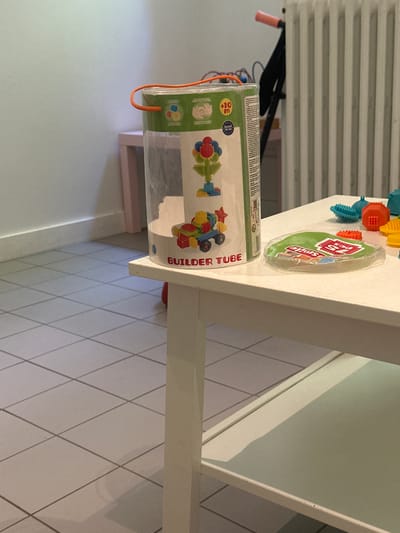 The image shows a close-up of a white table with some children's toys on it. The toys include a clear plastic tube filled with colorful building pieces and a green sticker with text. The text on the sticker reads "BUILDER TUBE". Other toys visible are scattered around the table and appear to be part of a building set. In the background, there is a white radiator and a pink chair. The lighting suggests it is daytime, and the scene appears to be indoors, likely in a home or play area. The overall impression is that of a child's play space.