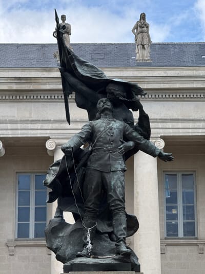 A bronze statue depicts a soldier with his arms outstretched, one holding a flagstaff and the other reaching out. He is leaning against a female allegorical figure. A white rope is tied around the soldier's legs. In the background, a building with columns and windows is visible, and two stone statues stand on the roof. The scene is set in Nantes, France, at the Place du Commerce, where the statue commemorates the sacrifices of the 85th Infantry Regiment during World War I.